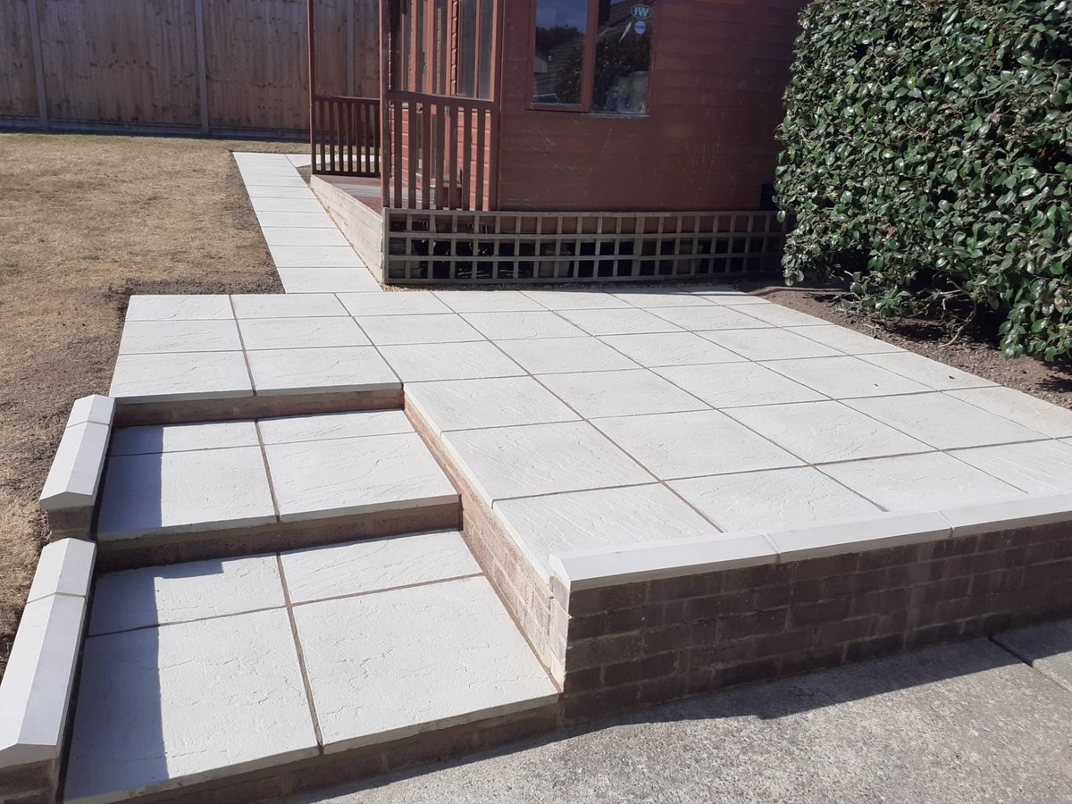 Modern patio with light-coloured tile pavers arranged in a geometric pattern, featuring brown brick edging and tiered steps leading up to a red brick building